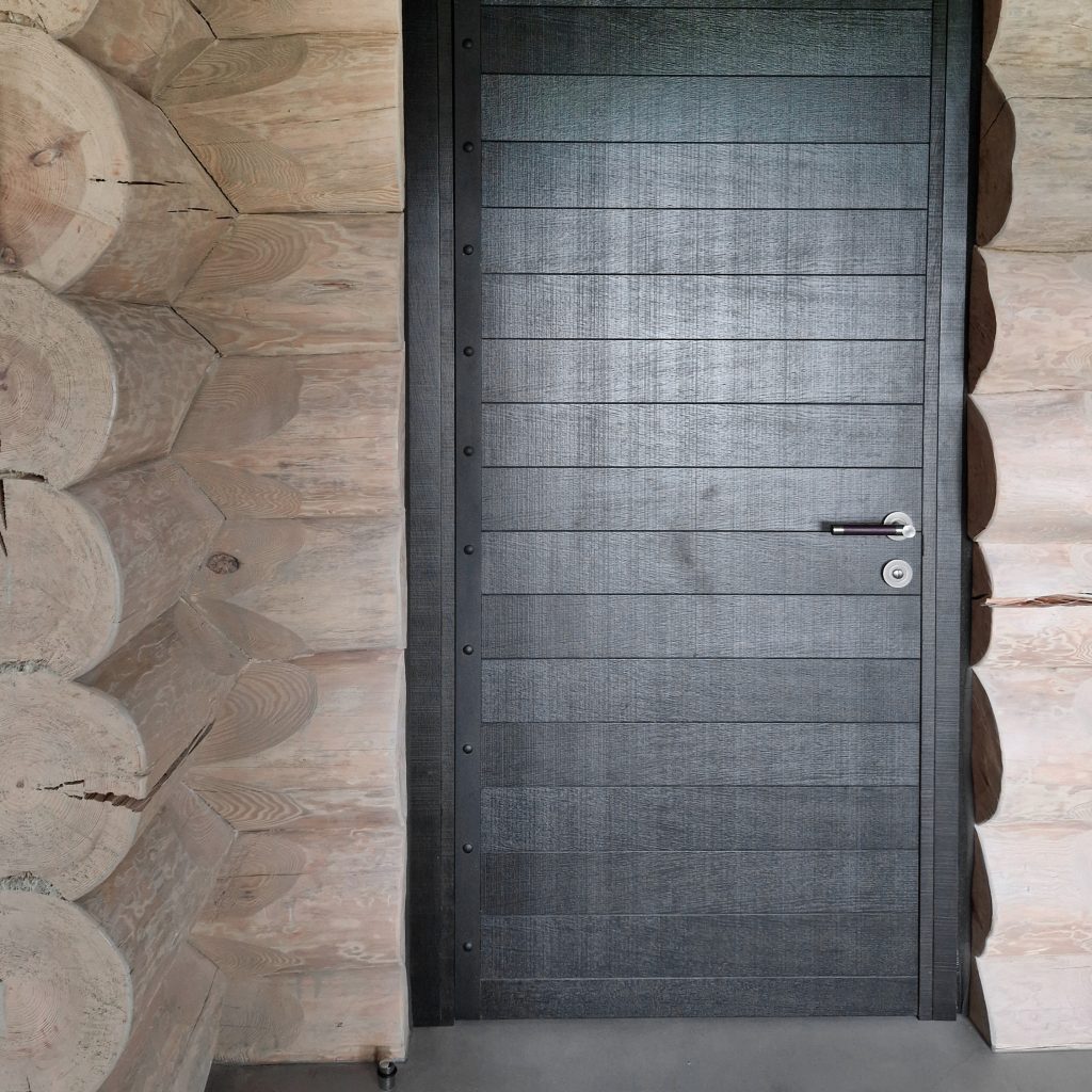 A wooden log walled corridor leads to a dark wooden door with Turnstyle Designs leather door lever has the handle. Natural materials have been chosen for this project to match the wooden walls, concrete floor and brass fixings.