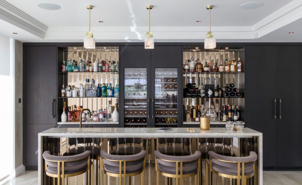 Luxury home bar with large marble island. This home bar based in the entertaining area of a large kitchen extension, has dark moody timber wood units on the back wall. Floating shelves either side of a large integrated wine cooler show off the owners large spirit collection and beautiful glassware. Four bar stools face the large marble island.