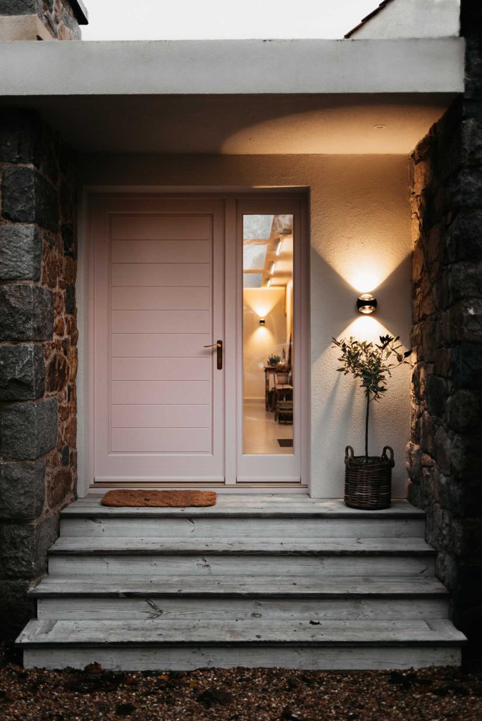 Grey stone steps and cobbled walls lead to a soft pink front door with warm orange lighting glowing from inside.