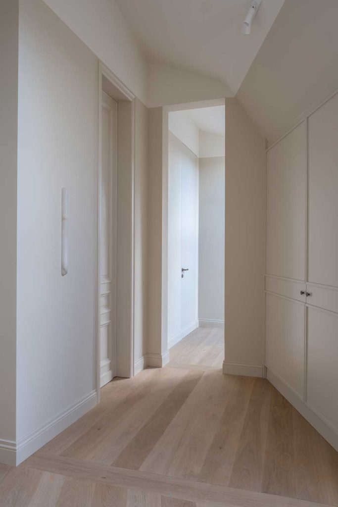 A corridor with character and angles has a neutral and clean finish of white doors and walls with oak parquet flooring. The doors and cupboards have Turnstyle Designs luxury door and furniture hardware on them.