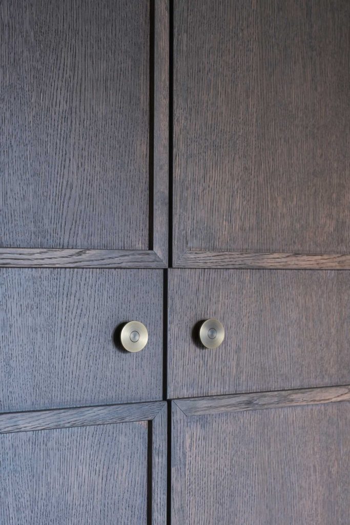 Decorative brass cabinet knobs by Turnstyle Designs have been used on modern panelled cupboard doors. The cabinet knobs are large flat circles. The cupboards doors are a dark brown wood with decorative panelling on each door.