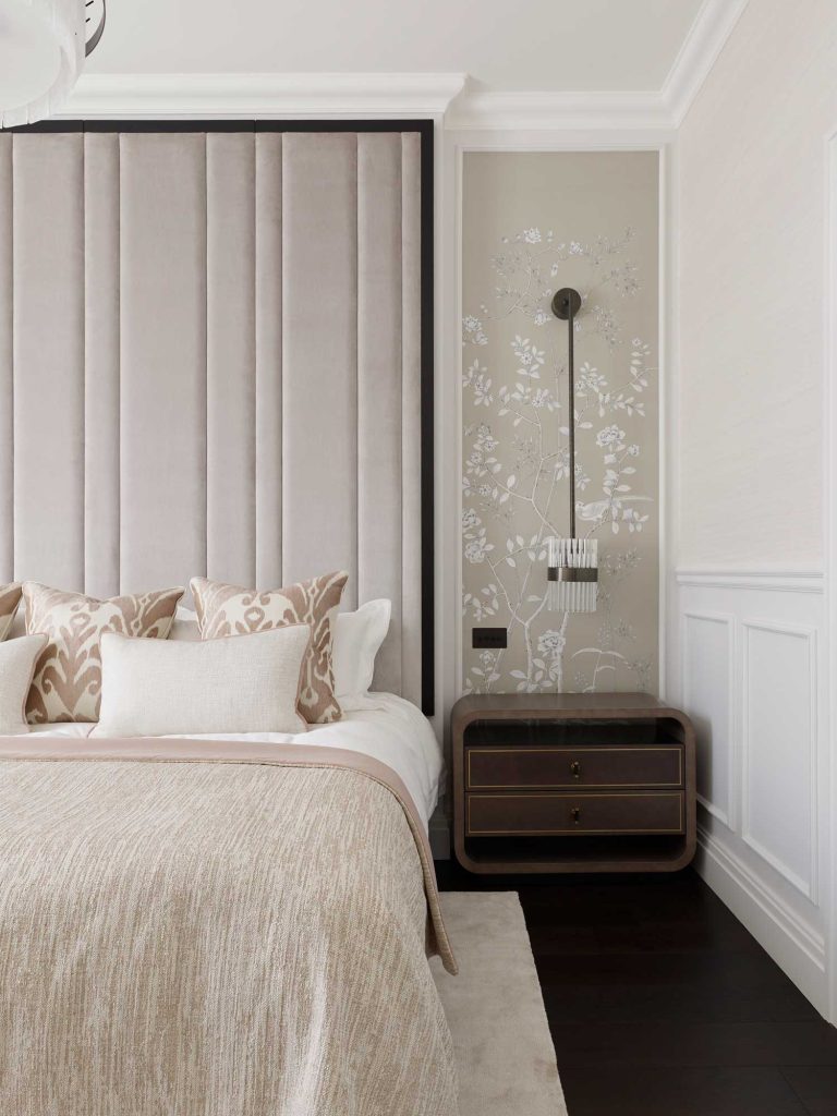 Luxury bedroom side table in deep brown wood with large headboard that reaches to the ceiling. Neutral and warm colours have been used for soft furnishings and decorative motif wallpaper to the side of the bed.