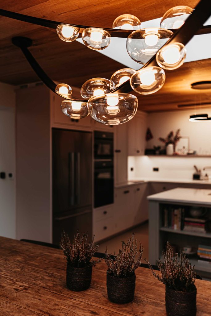Moody draped lighting sits over a family sized wooden table in a modern and clean kitchen.