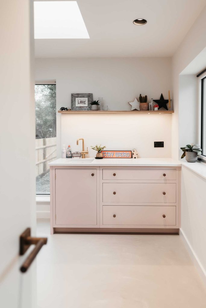 Looking it a side room of an open plan kitchen, this modern bright kitchen has soft pink units and white counter tops. The contrasting deep brass hammered cabinet knobs are made by Turnstyle Designs.