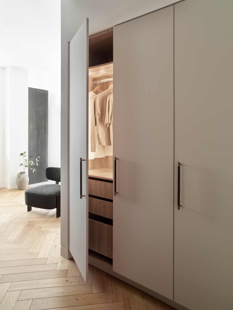 Large built in wardrobe with Turnstyle Designs handstitched leather pull handles. The neutrally painted wardrobe sits on parquet flooring within a master bedroom set up. The wardrobe doors are slightly ajar showing inside lighting.