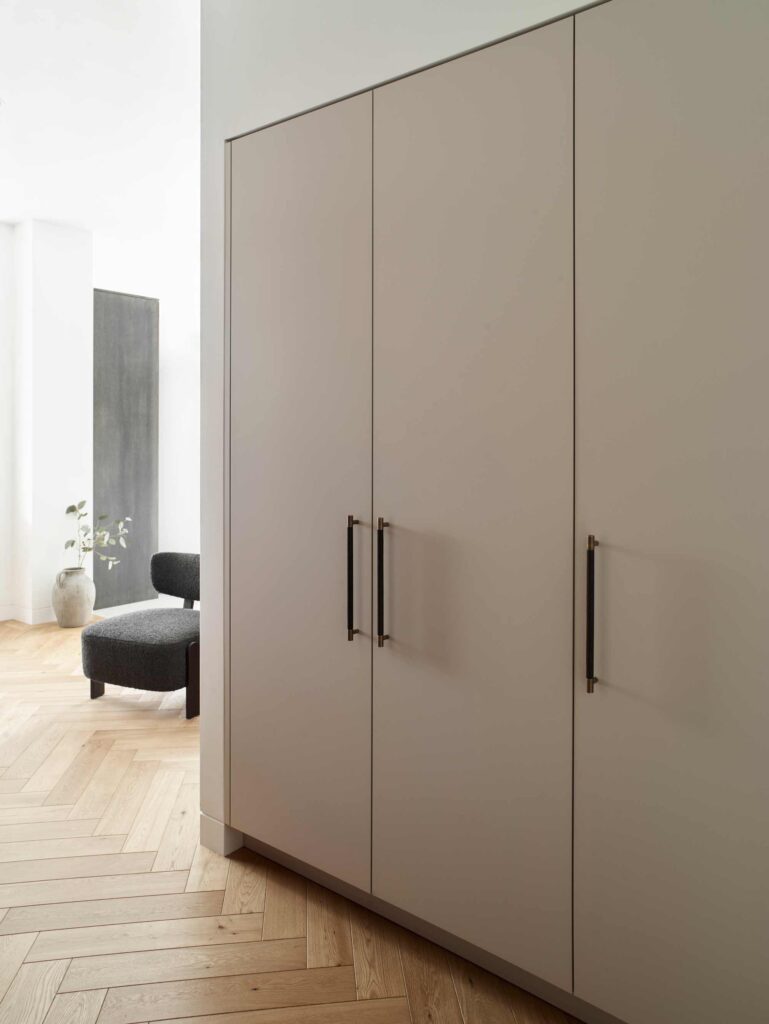 Large built in wardrobe with Turnstyle Designs handstitched leather pull handles. The neutrally painted wardrobe sits on parquet flooring within a master bedroom set up.